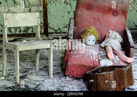 Salle de jeux dans un bâtiment de Prypiat. Une poupée décapitée est posée sur une chaise rouge brisée à côté d'un siège vide. Zone d'exclusion de Tchernobyl, Ukraine Banque D'Images