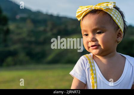 gros plan d'une belle petite fille latina à la peau brune, portée par sa mère tout en regardant le sol, portant une robe jaune sur un beau Banque D'Images