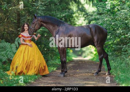 Une jeune femme vêtue d'une robe jaune vintage marche avec un cheval brun dans un parc vert le jour de l'été Banque D'Images