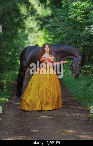 Une jeune femme vêtue d'une robe jaune vintage marche avec un cheval brun dans un parc vert le jour de l'été Banque D'Images