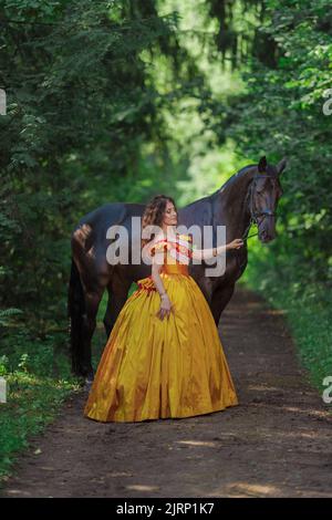 Une jeune femme vêtue d'une robe jaune vintage marche avec un cheval brun dans un parc vert le jour de l'été Banque D'Images
