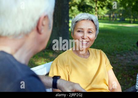 Beau couple plus âgé qui parle assis sur le banc dans le parc d'été, porter des vêtements de sport, profiter de la conversation le matin à l'extérieur. Communication, préjudice Banque D'Images