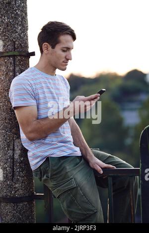 Un adolescent assis sur la main courante à l'extérieur et tapant des messages sur le téléphone. Banque D'Images