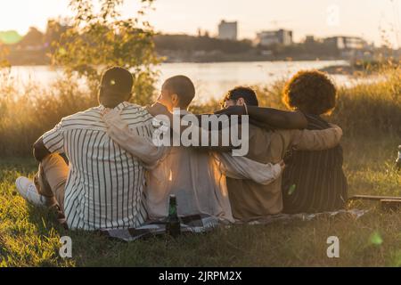 Vue arrière de multiculturel bon vieux amis qui s'embrasse tout en étant assis sur une couverture sur l'herbe, ayant un pique-nique de fin de semaine dans la nature près de la rivière, parler, boire de la bière et admirer la vue. Heure du coucher du soleil. Photo de haute qualité Banque D'Images