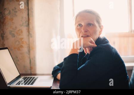 Femme mûre réfléchie assise près d'un ordinateur portable travaillant à la maison Banque D'Images