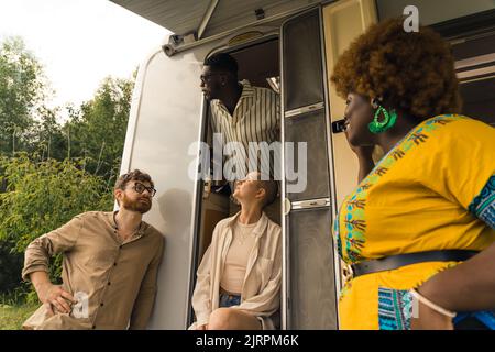 Voyage en camping. Un groupe multiculturel de quatre bons amis se tenant près de leur confortable motorhome et assis sur des marches pliantes tout en parlant, en riant et en appréciant passer du temps ensemble dans la nature pendant les vacances d'été . Photo de haute qualité Banque D'Images