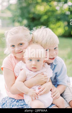 Deux enfants blonds embrassent leur bébé sœur aux yeux bleus dans le parc. Banque D'Images
