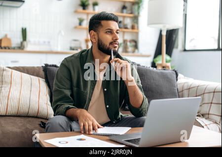 Un homme indien ou arabe pensif, étudiant ou indépendant, utilisant un ordinateur portable, travaillant ou étudiant à distance, écoutant des webinaires, prenant des notes, devoirs, assis à la maison sur un canapé, regarde, pense Banque D'Images