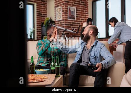 Une équipe diversifiée d'amis faisant le geste de HighFive après avoir gagné des jeux vidéo jouer sur la console de télévision. Célébrez la victoire de la compétition ensemble lors de la fête à la maison et de la rencontre avec les gens. Banque D'Images