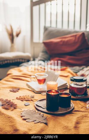 Livre ouvert, bougies en feu et tisane dans une tasse de verre sur le écossais jaune sur le lit dans la chambre confortable. Lecture, détente, loisirs à la maison. Message d'humeur automatique. Banque D'Images