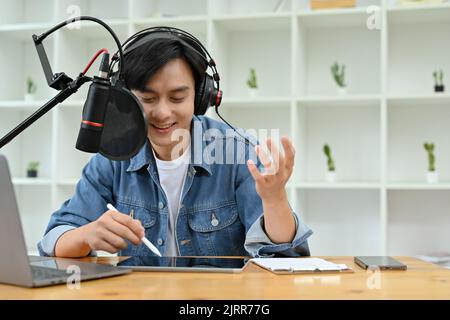 Homme asiatique gai dans un casque et microphone professionnel enregistrant un podcast audio pour la radio, chaîne en direct sur Internet en studio. Banque D'Images