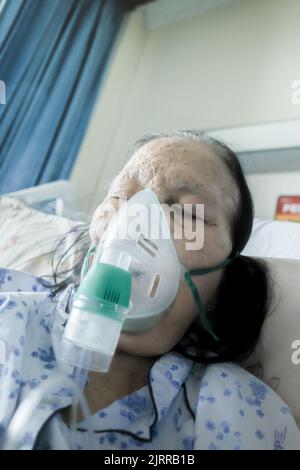 Patiente âgée d'Asie du Sud-est hospitalisée et sous traitement respiratoire par nébuliseur dans un lit d'hôpital Banque D'Images