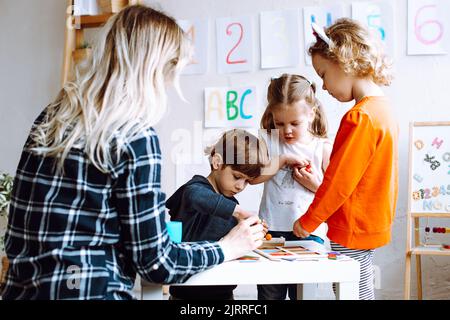 Deux jolies petites filles debout à table. Petit garçon jouant à des jeux de société. Jeune femme enseignante aidant les enfants à jouer. Banque D'Images