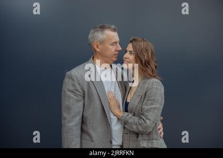 Jeune femme aimante et beau partenaire âgé gris à cheveux embrassant et regardant l'un l'autre, fond gris. Relations romantiques de beau couple Banque D'Images