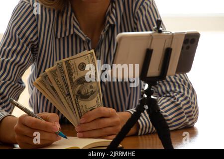 Une fille avec des dollars dans ses mains parle sur un chat vidéo au téléphone à une table dans le bureau, un appel en ligne pour le travail, la finance et le succès, un sem Banque D'Images