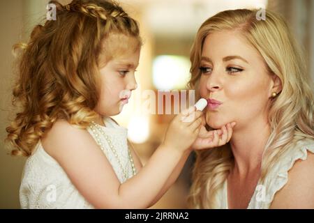 Et un peu de rouge à lèvres pour la reine aussi... une petite fille appliquant le rouge à lèvres à sa mère à la maison. Banque D'Images