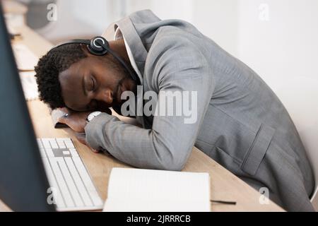 Un agent du centre d'appels CRM fatigué dormant sur le bureau de l'ordinateur au bureau avec un casque allumé. Un jeune homme d'affaires épuisé prend une sieste sur la table. Africain Banque D'Images