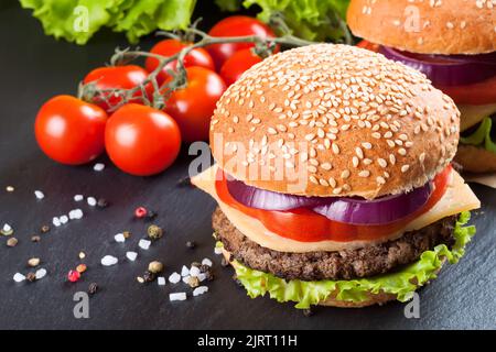 Cheeseburger maison avec viandes de bœuf, salade fraîche, tomates et oignons sur des petits pains à l'assaisonnement, servis sur une table en ardoise noire. Banque D'Images