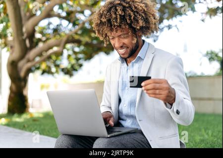 Homme brésilien ou hispanique positif, assis à l'extérieur, en utilisant une carte bancaire et un ordinateur portable, faire des achats en ligne, faire des paiements, commander des livraisons de nourriture, entrer des données, sourire. Achats en ligne, paiement Banque D'Images