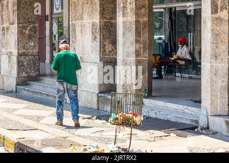 Un homme cubain marche à côté d'une poubelle avec la moitié des ordures dans le sol. En arrière-plan, un autre homme est assis dans une chaise sous le porche Banque D'Images