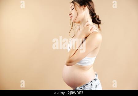 Vue latérale d'une femme enceinte souffrant de douleurs dans le bas du dos, fond beige. Banque D'Images