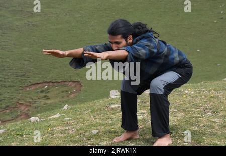 Un jeune Indien long poil pratiquant la pose d'ours (chaise pose variation Forward Stretch) en extérieur. Un jeune homme faisant Utkatasana yoga pose (variation Forward Stretch). Banque D'Images
