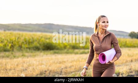 Belle femme marchant dans le champ avec tapis de yoga en main avec grand sourire à la recherche d'un endroit pour les loisirs Banque D'Images