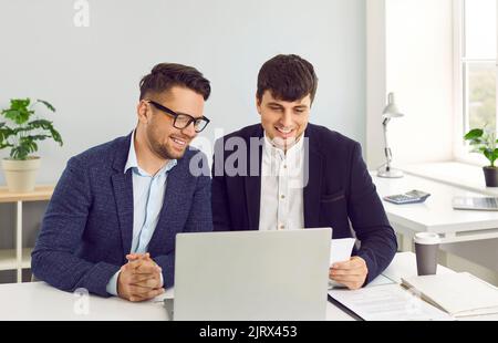 Deux jeunes hommes d'affaires heureux travaillant au bureau et utilisant un ordinateur portable ensemble Banque D'Images