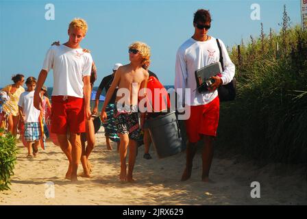 Un jeune garçon marche et parle à deux sauveteurs en quittant la plage, portant leur équipement lors d'une journée de vacances d'été à la plage Banque D'Images