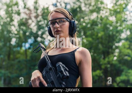 Gros plan de taille moyenne d'une belle fille de race blanche, douée et confiante, regardant un appareil photo, portant des lunettes de protection et des écouteurs, et tenant une carabine noire. Arrière-plan flou. Prise de vue en extérieur. Photo de haute qualité Banque D'Images