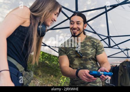 Distributeur d'armes à feu à poil sombre caucasien dans un t-shirt moro montrant une arme à feu bleu foncé à sa cliente à poil long avec un sourire sur son visage. Photo de haute qualité Banque D'Images