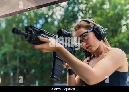 Jeune femme européenne adulte ciblée regardant à travers la carabine et se préparant à une compétition au champ de tir. Arbres flous en arrière-plan. Prise de vue en extérieur. Photo de haute qualité Banque D'Images