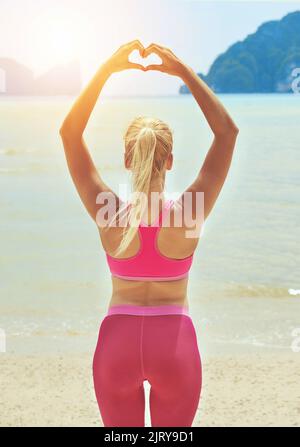 Tombez amoureux de votre entraînement. Vue arrière d'une jeune femme sportive en forme de coeur avec ses mains, debout sur la plage. Banque D'Images