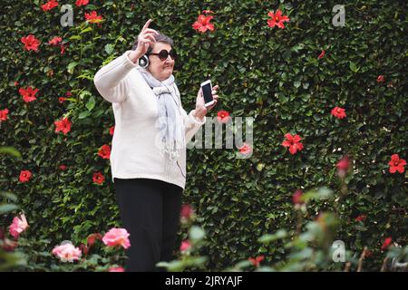 Drôle vieille femme dansant écouter de la musique sur un smartphone portant des écouteurs souriant en appréciant le plaisir de célébrer la retraite dans le jardin avec mur de fleurs Banque D'Images