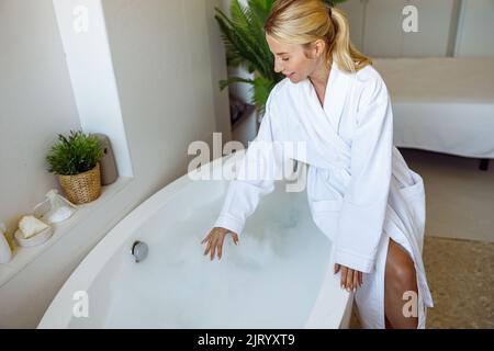 Vue d'en haut sur une jeune femme blonde assise dans la salle de bains et préparant le bain avec des bulles. Banque D'Images