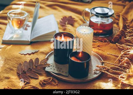 Automne confortable à la maison. Bougies allumées avec carte hello automne, tisane dans verre tasse et livre ouvert sur le écossais jaune. Lecture, détente, loisirs à Banque D'Images