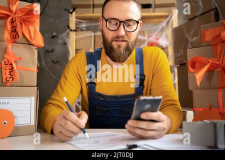 Concept de PME. Beau jeune liveur heureux avec la nouvelle commande du client, écrivant des boîtes en utilisant le téléphone intelligent dans le bureau à domicile, propriétaire de petite entreprise Banque D'Images