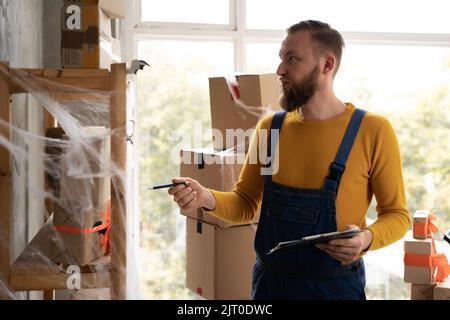 jeune homme barbu en lunettes propriétaire d'affaires assis dans halloween décoré bureau à la maison. Plie les boîtes avec des noeuds orange. Vend des marchandises pour les vacances de tous Banque D'Images