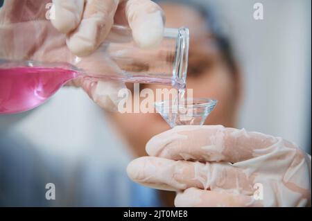 Gros plan mains gantées d'un chimiste scientifique qui verse une substance chimique liquide rose dans un flacon en verre à travers un entonnoir Banque D'Images