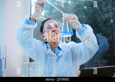 Femme afro-américaine, chimiste scientifique qui goutte peu de réactif dans une fiole à fond plat avec une solution chimique bleue Banque D'Images