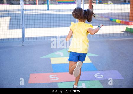 Une vue arrière d'une fille avec une chemise jaune et des queues de merlu jouant le hopscotch dans l'aire de jeux Banque D'Images