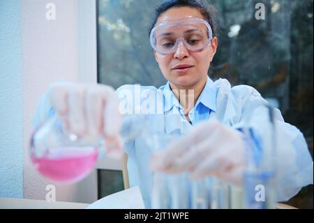 Une femme afro-américaine, pharmacienne pharmacologue, verse la substance liquide rose d'une fiole de laboratoire en verre dans des tubes à essai Banque D'Images