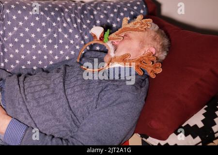 Homme de 50-60 ans dormant sur un canapé portant une décoration sous forme de cornes de cerf sur la tête. Homme reposant après Noël et fête du nouvel an. Banque D'Images