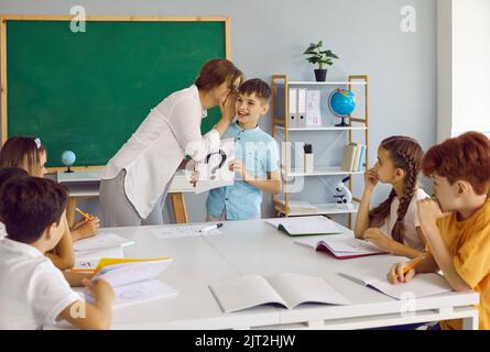 Une enseignante amicale murmure à l'oreille d'un petit garçon donnant un indice pendant la leçon scolaire. Banque D'Images