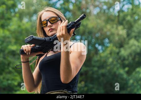 Concept de plage de prise de vue en extérieur. Jeune belle fille caucasienne à cheveux longs dans un haut de réservoir noir regardant de près un fusil noir. Arbres flous en arrière-plan. Prise de vue moyenne. Photo de haute qualité Banque D'Images