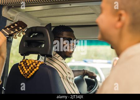 Groupe multi-ethnique d'amis proches ayant un voyage d'été dans une camionnette de camping, conduite, jouer de la guitare, parler et rire chaleureusement à l'intérieur de leur véhicule. Le conducteur regardant ses copains. Photo de haute qualité Banque D'Images