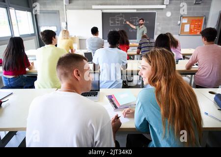L'enseignant explique la leçon sur le tableau noir en arrière-plan tandis qu'en premier plan deux élèves parlent l'un à l'autre. Camarades de classe du lycée Banque D'Images