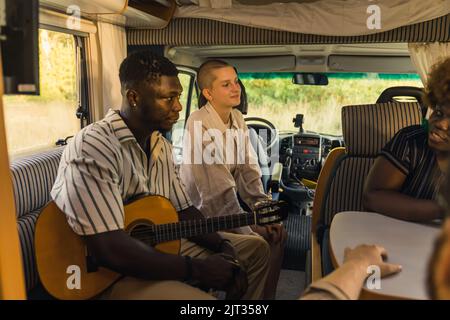 Groupe multi-ethnique d'amis proches lors d'un voyage d'été assis à l'intérieur de leur confortable motorhome moderne, jouant de la guitare, parlant et riant. Photo de haute qualité Banque D'Images