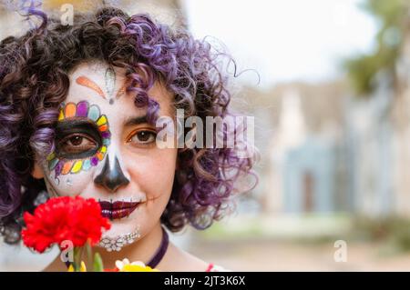 Portrait de belle jeune femme latine à l'extérieur regardant l'appareil photo et souriant, tenant une fleur rouge et portant la Calavera Catrina maquillage, copie spac Banque D'Images