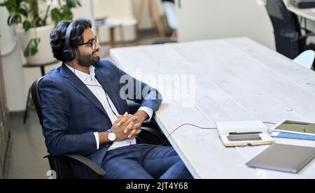 Un jeune homme d'affaires indien portant des écouteurs assis au travail écoute podcast. Banque D'Images